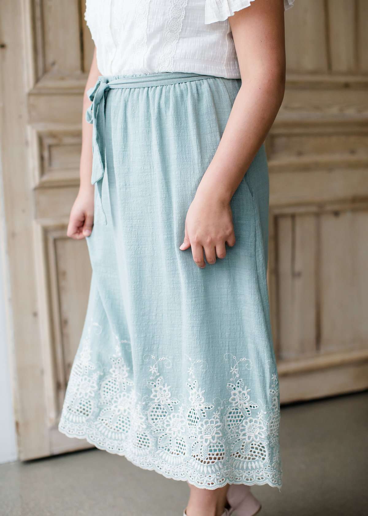 Woman wearing a mint green cotton midi skirt with a self tie and feminine crochet lace detail at the bottom hem