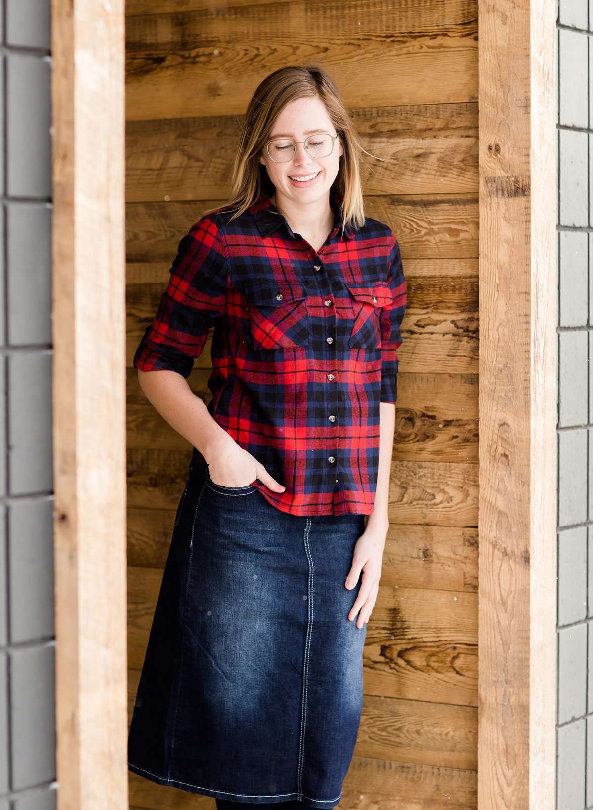 Woman wearing a cozy, checkered plaid top. This button up plaid shirt features roll tab sleeves and is black, navy and red. It is also paired with a below the knee jean skirt.