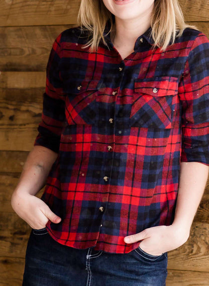 Woman wearing a cozy, checkered plaid top. This button up plaid shirt features roll tab sleeves and is black, navy and red. It is also paired with a below the knee jean skirt.