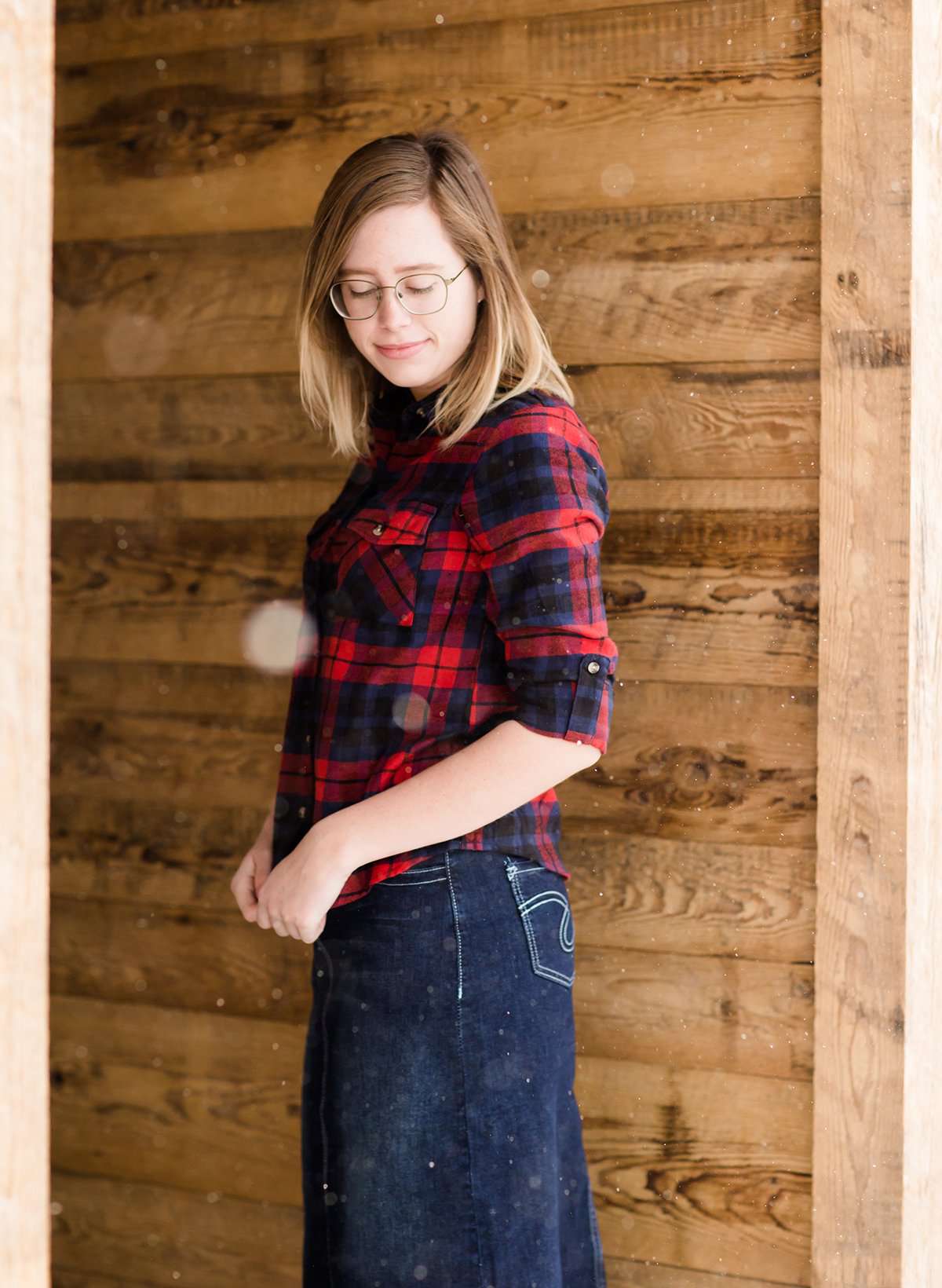 Woman wearing a cozy, checkered plaid top. This button up plaid shirt features roll tab sleeves and is black, navy and red. It is also paired with a below the knee jean skirt.