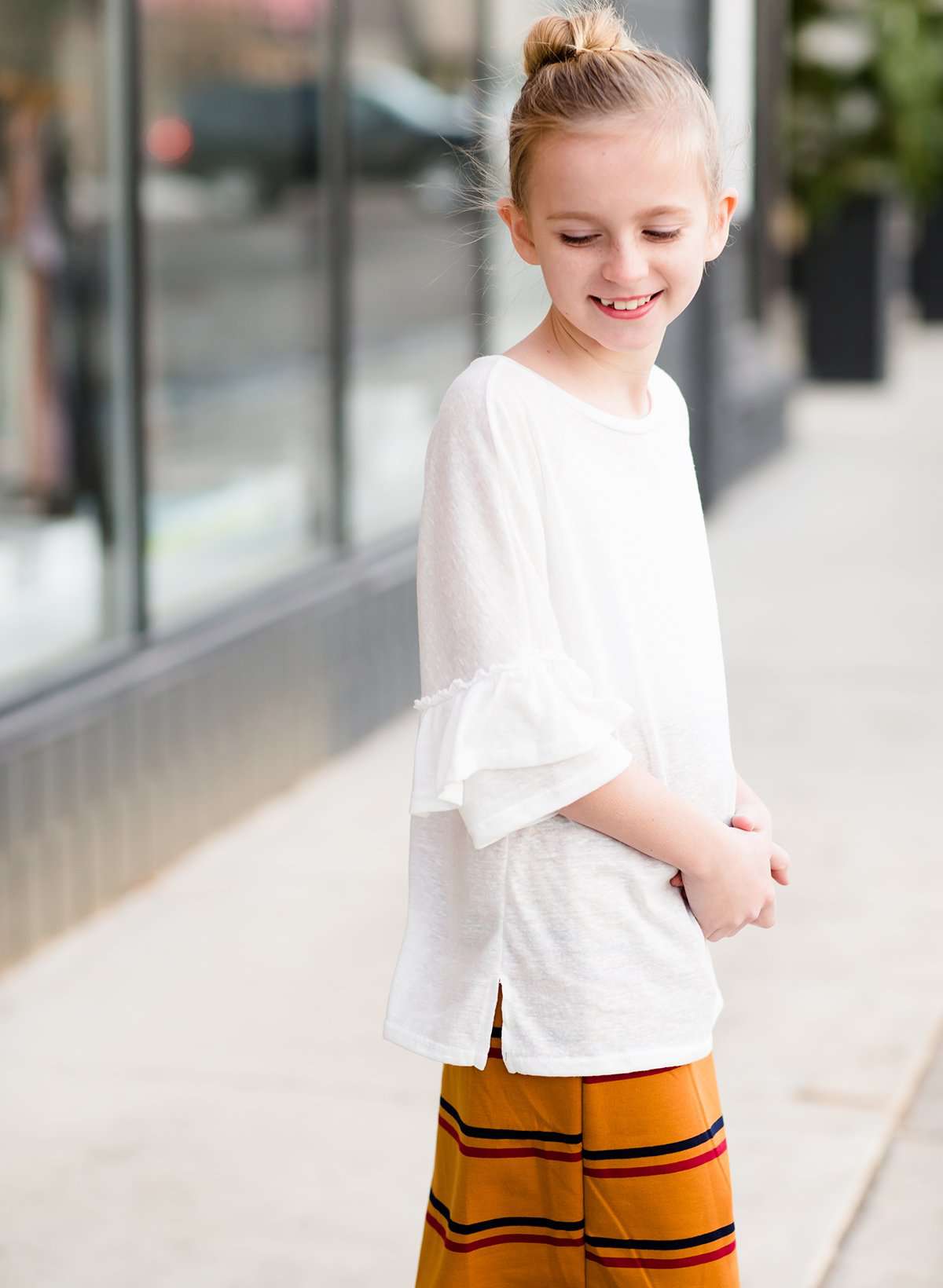 Young girl wearing an ivory, oversized modest top with ruffle sleeve accents. This top is also paired with a mustard, striped below the knee skirt and short boots.