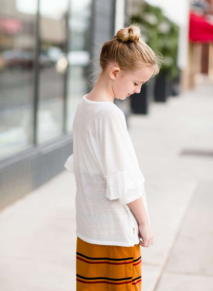 Young girl wearing an ivory, oversized modest top with ruffle sleeve accents. This top is also paired with a mustard, striped below the knee skirt and short boots.