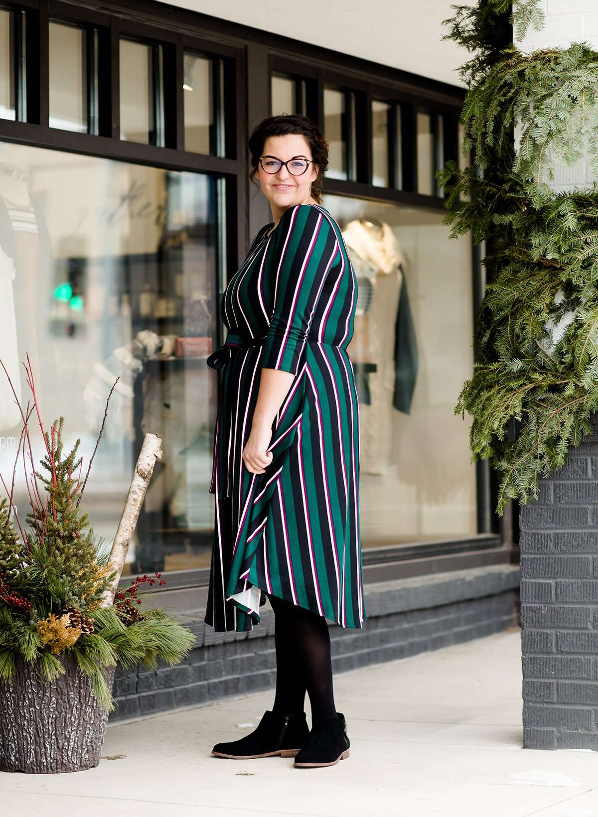 Modest woman wearing a green and black striped midi dress with a self tie bow. She is wearing black leggings and black booties as well. She is at Inherit Clothing Company.