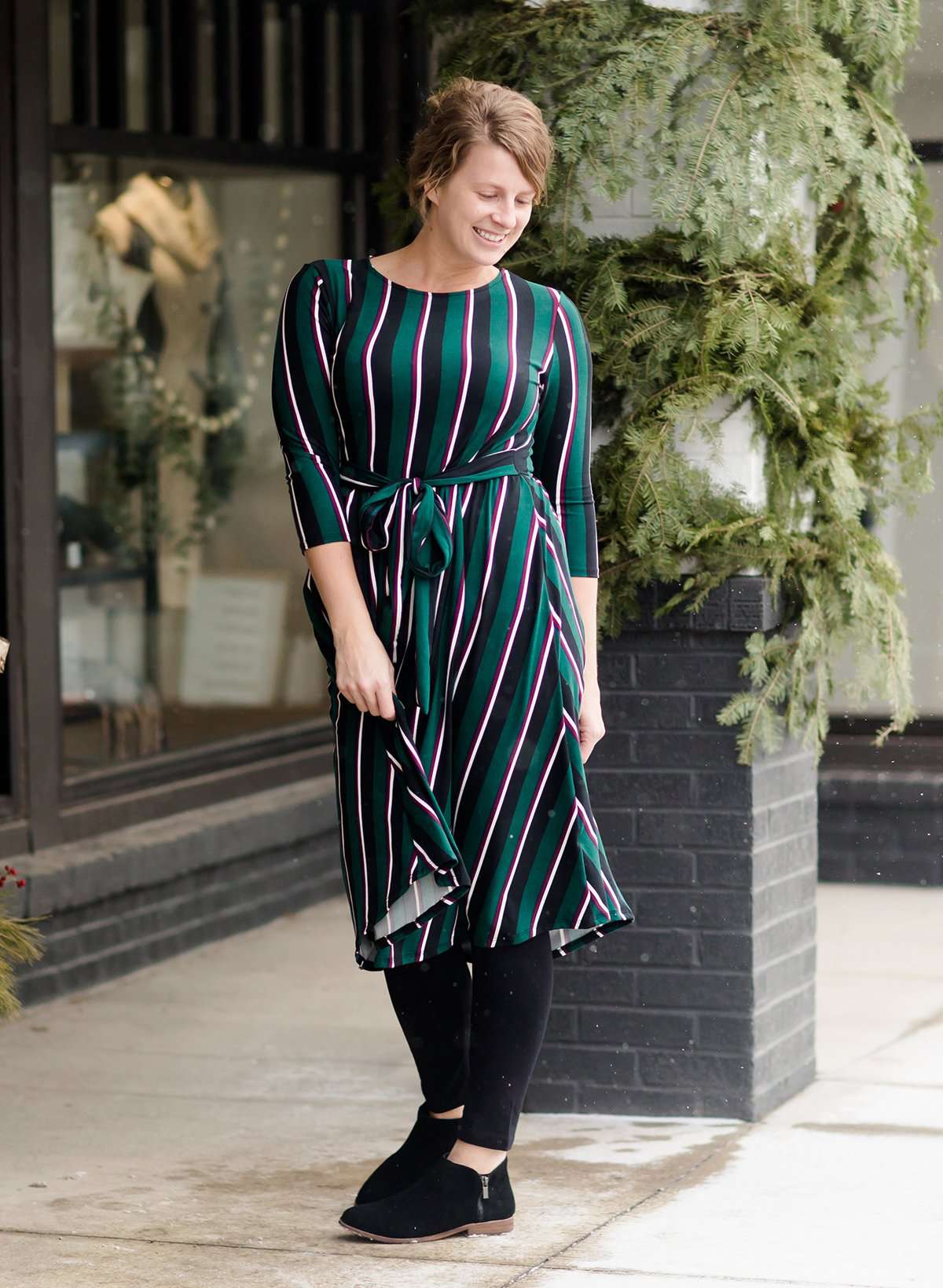 Modest woman wearing a green and black striped midi dress with a self tie bow. She is wearing black leggings and black booties as well. She is at Inherit Clothing Company.