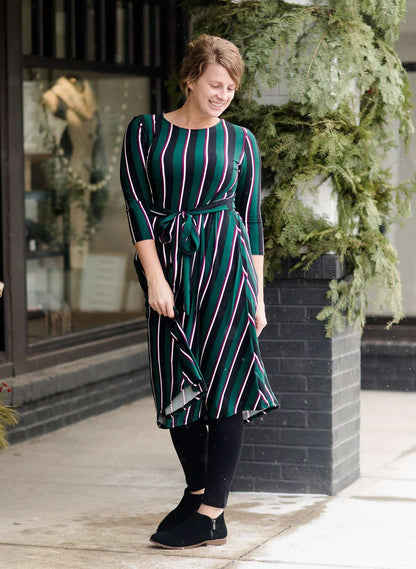 Modest woman wearing a green and black striped midi dress with a self tie bow. She is wearing black leggings and black booties as well. She is at Inherit Clothing Company.