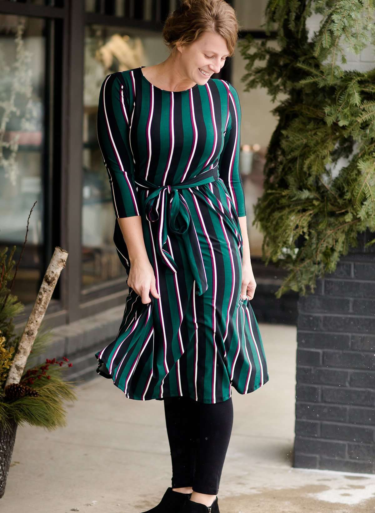 Modest woman wearing a green and black striped midi dress with a self tie bow. She is wearing black leggings and black booties as well. She is at Inherit Clothing Company.