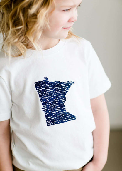 Toddler girl wearing a white minnesota logo on a white tee shirt. She is also wearing a long khaki skirt.