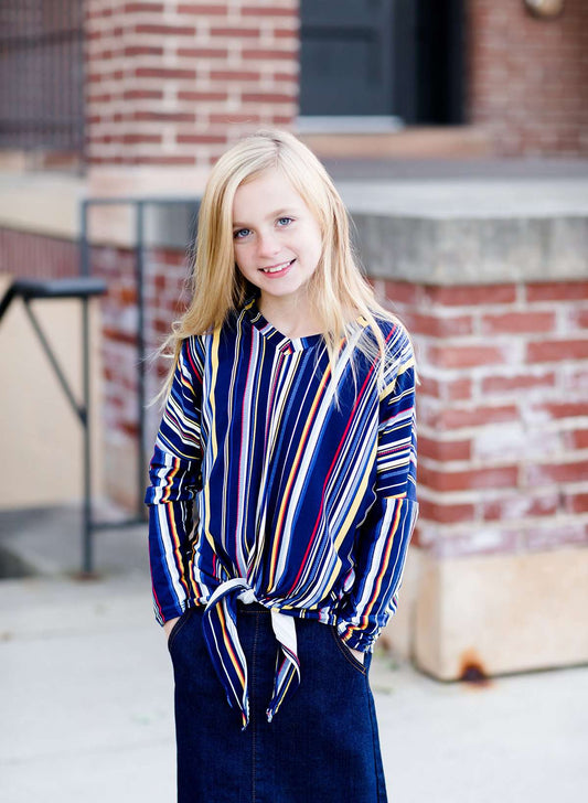 Young girl wearing a multi colored striped dolman top with a front tie. Colors in this top are navy, yellow, red, purple, green and white!