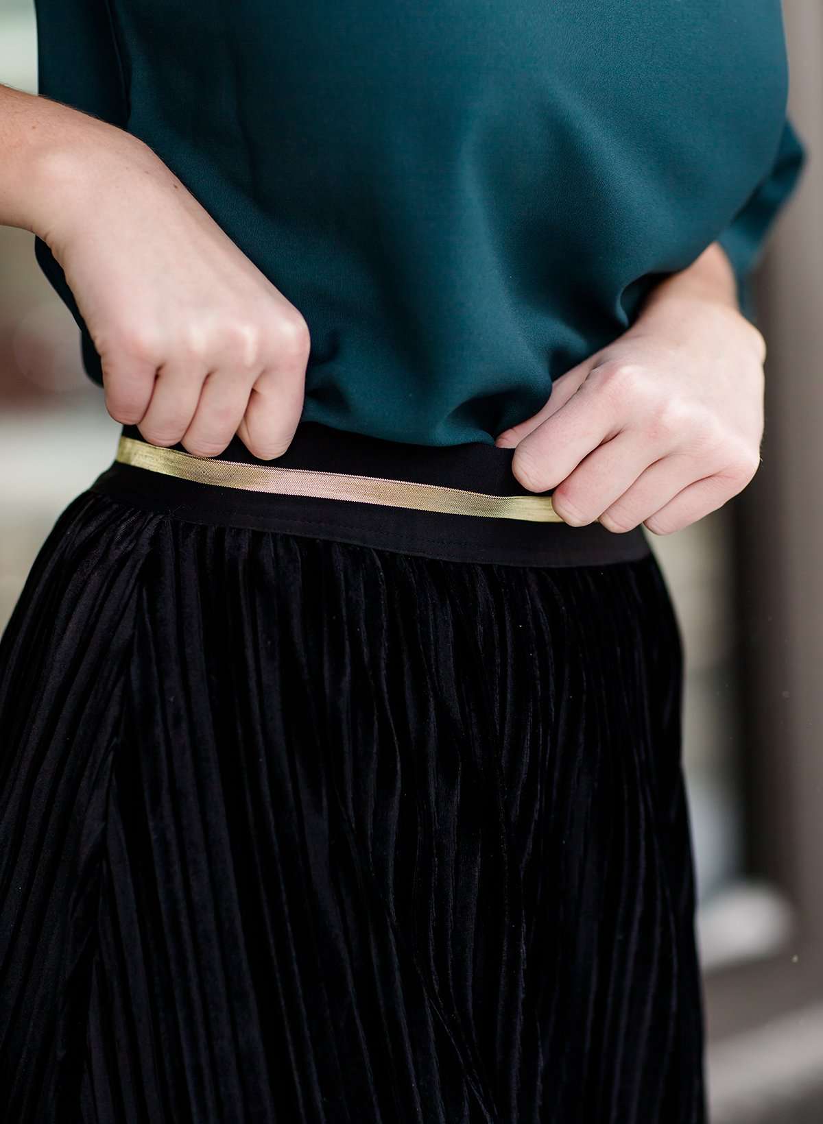 Woman wearing a black, velvet pleated midi skirt. This skirt has a gold accent around the elastic waistband. This modest skirt is paired with a teal, ruffle sleeve poets blouse.