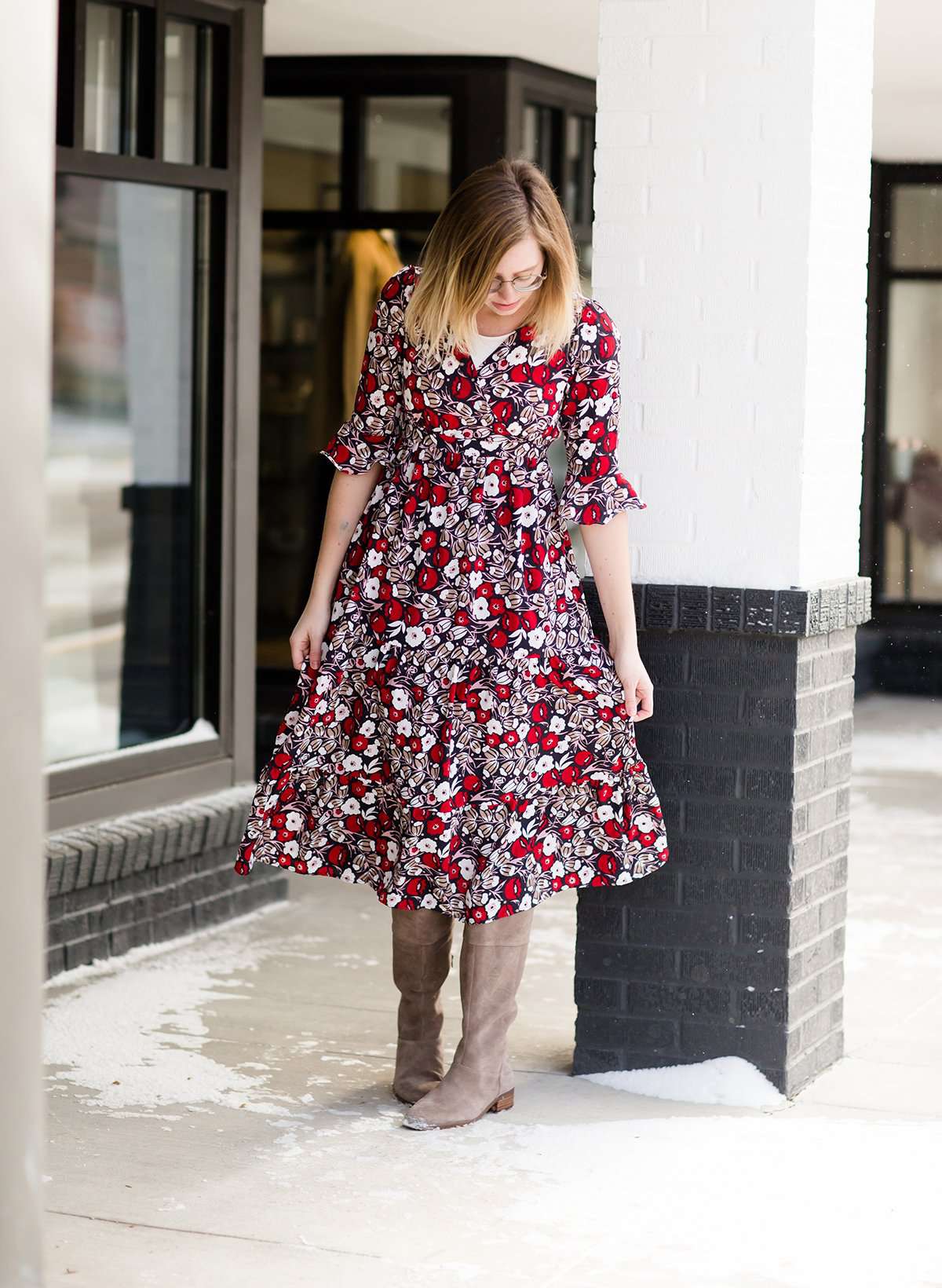 Woman wearing a midi length modest dress. This dress is a holiday red with poppy flowers and white flowers on it. It has a v-neck and ruffle 3/4 sleeves. This dress is paired with tall suede riding boots.