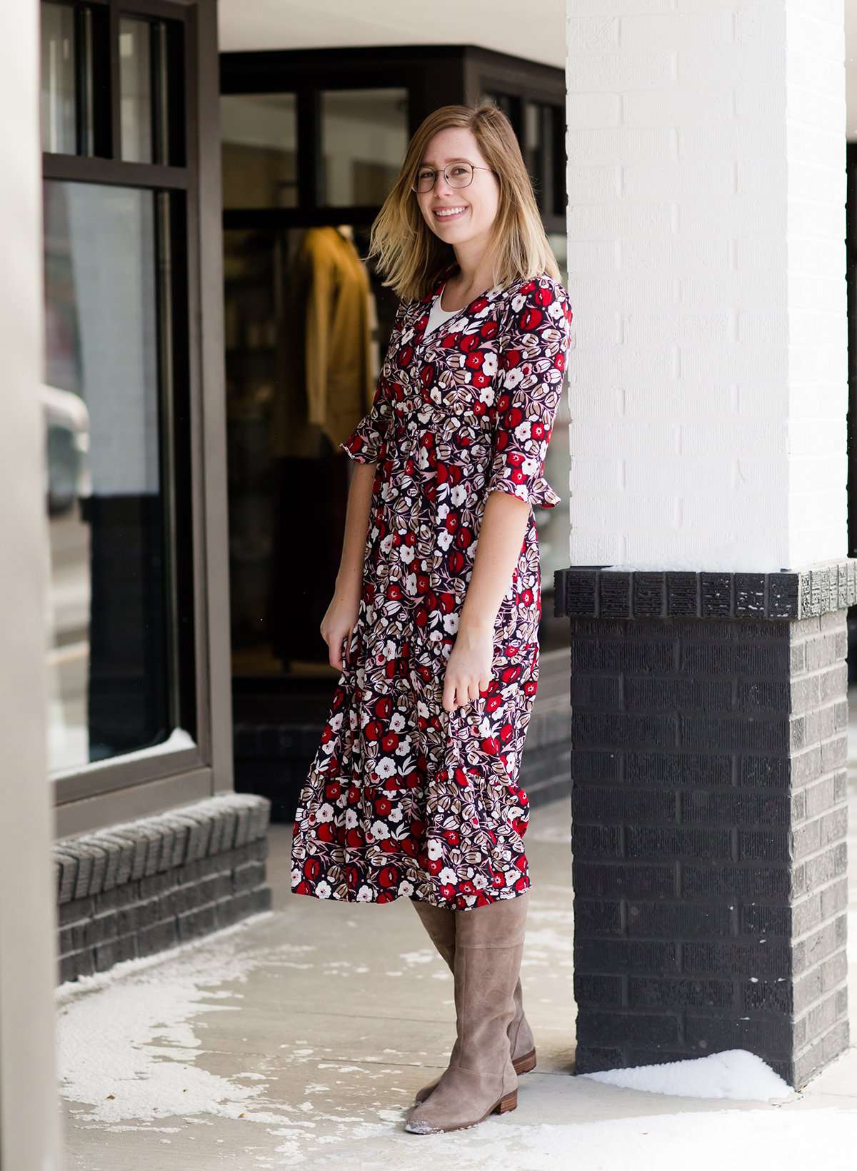 Woman wearing a midi length modest dress. This dress is a holiday red with poppy flowers and white flowers on it. It has a v-neck and ruffle 3/4 sleeves. This dress is paired with tall suede riding boots.