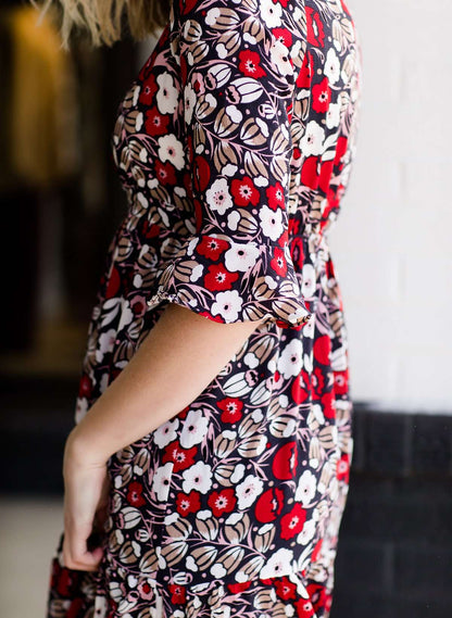 Woman wearing a midi length modest dress. This dress is a holiday red with poppy flowers and white flowers on it. It has a v-neck and ruffle 3/4 sleeves. This dress is paired with tall suede riding boots.