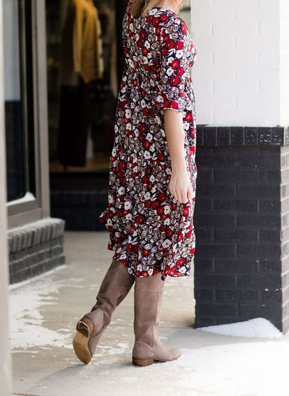 Woman wearing a midi length modest dress. This dress is a holiday red with poppy flowers and white flowers on it. It has a v-neck and ruffle 3/4 sleeves. This dress is paired with tall suede riding boots.